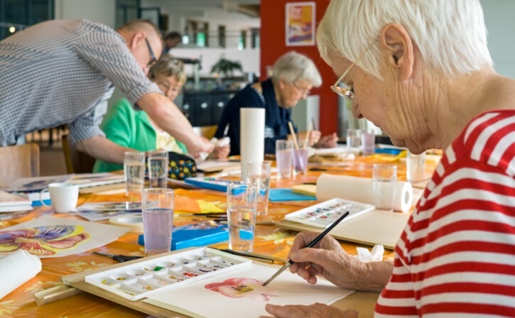 Woman in striped red and white shirt working on watercolor painting at table with other students in spacious studio.