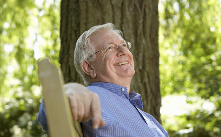 Senior man sitting on garden bench, looking up, smiling, close-up